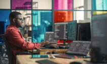 African American Man Writing Lines Of Code On Desktop Computer With Multiple Monitors and Laptop in Creative Office. Male Data Scientist Working on Innovative Online Service For Start-up Company.